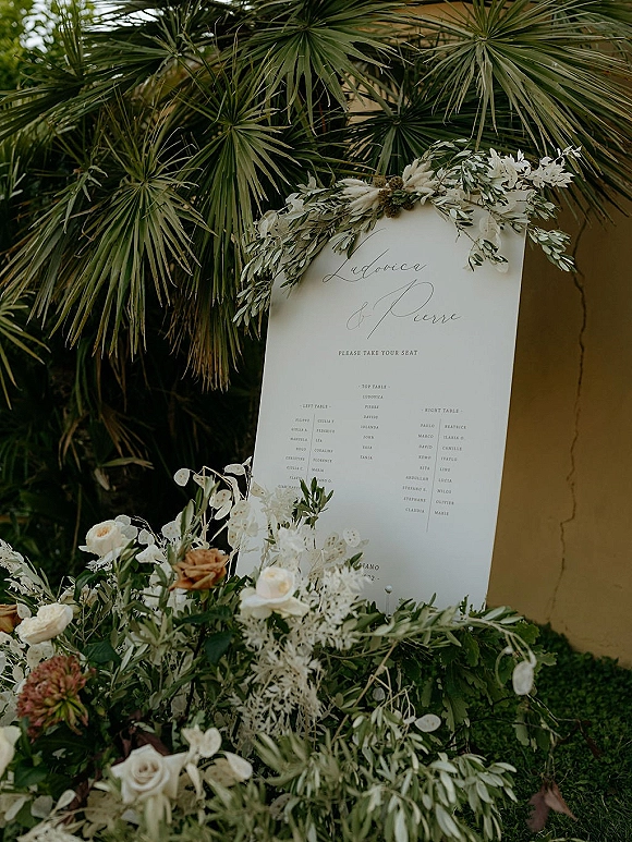 Wedding seating chart sign with calligraphy lettering, accented by white roses and greenery on an easel against a stucco wall outdoors