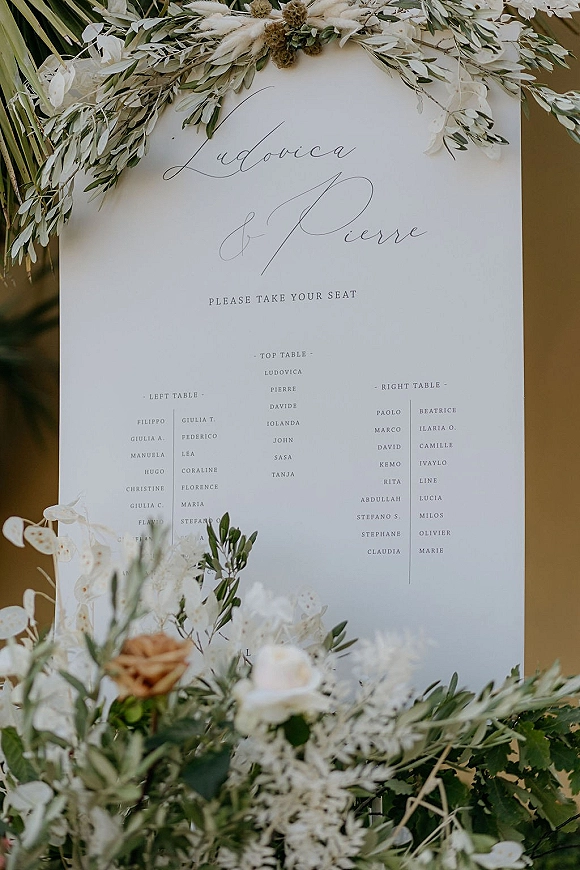 Wedding seating chart sign with elegant calligraphy lettering on a white board, styled with olive branches and dried florals against a neutral backdrop