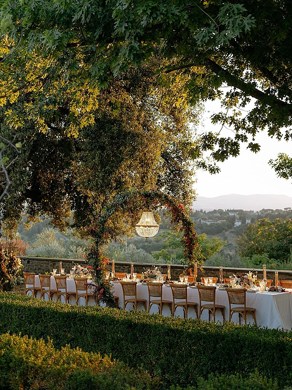 Outdoor reception tablescape with a long banquet table wedding setup, wicker chairs, floral arch and chandelier under trees by a stone wall