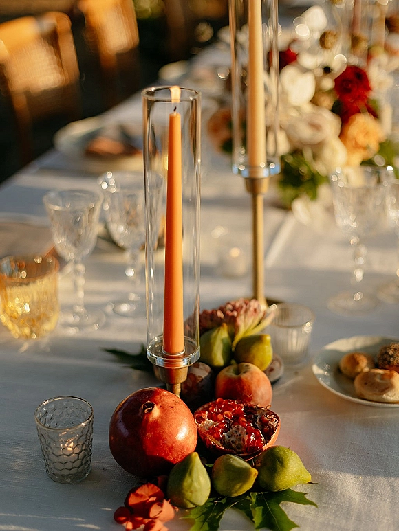 Reception tablescape with wedding table centerpiece of taper candles in glass hurricanes, brass sticks, red-white florals, and fruit on white linen outdoors in warm sunlight