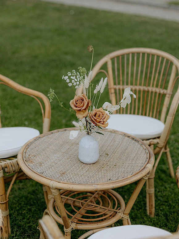 Cocktail table decor with a bud vase centerpiece of roses, wildflowers, and dried grasses on a wicker table beside rattan chairs on grass