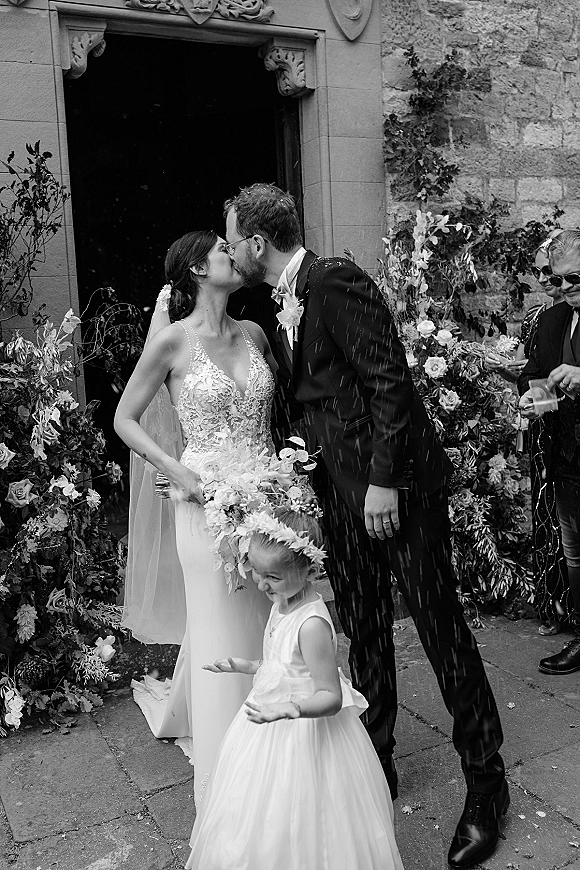 Wedding kiss as bride and groom embrace outside an ivy-covered stone church doorway, confetti falling as guests cheer behind them