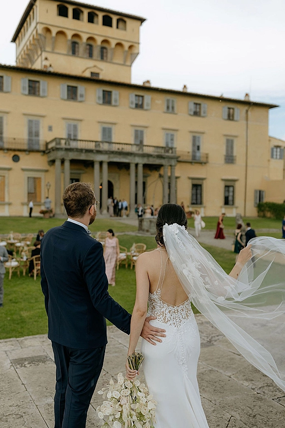 Wedding couple portrait from behind, bride holding bouquet with veil blowing as they walk on a stone terrace at a historic villa