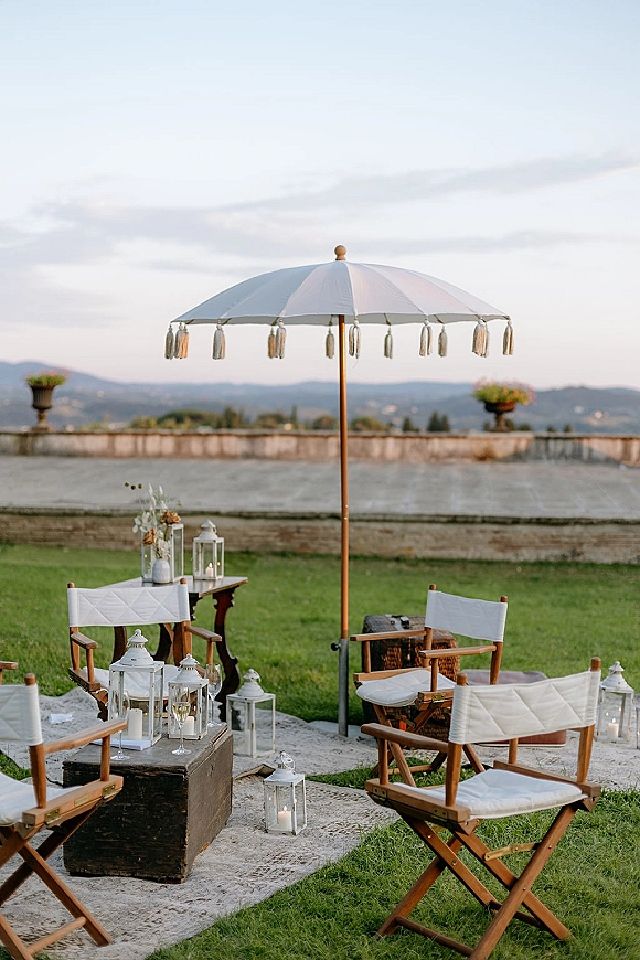 Wedding lounge seating with white tassel umbrella, director chairs, layered rugs, lanterns, candles and champagne flutes on a lawn terrace