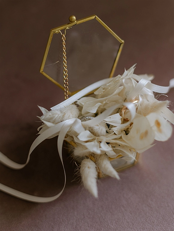 Wedding rings in a gold geometric glass ring box with a chain, styled with white dried florals and ribbon on a brown tabletop