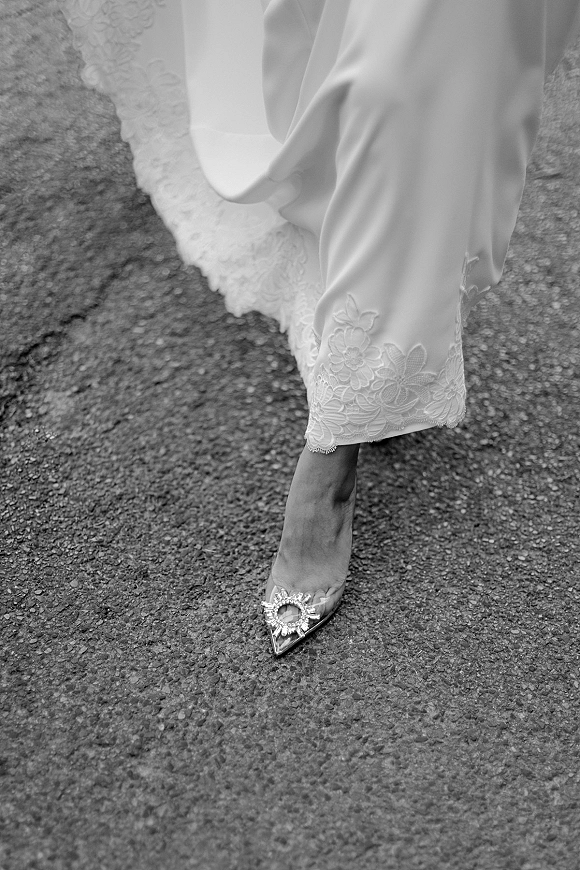 Bridal shoe detail showing a pointed toe heel with jeweled buckle beside lace wedding dress hem on asphalt pavement, black and white