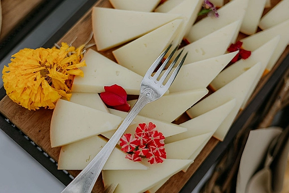 Wedding cheese board with pre-sliced cheese, serving fork, and edible flower petals arranged on a wooden tray over a rustic wood surface