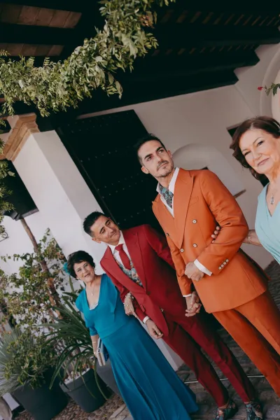 Family wedding portrait featuring a two-grooms portrait with their mothers, in orange and red suits by a white stucco archway with greenery garland.