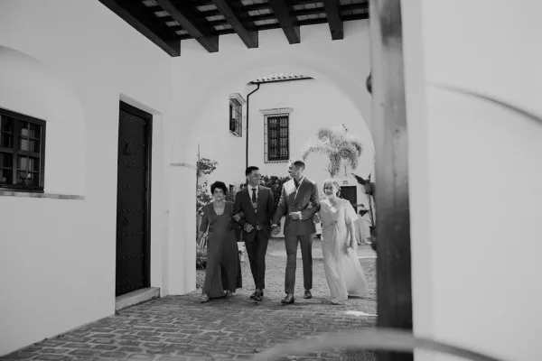 Wedding party candid in a black and white wedding photo, walking under white stucco arches in a cobblestone courtyard, one in a double-breasted suit.