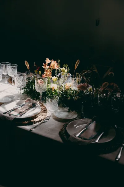 Reception tablescape for a wedding table setting, anchored by a floral centerpiece against a dark wall with soft shadows.