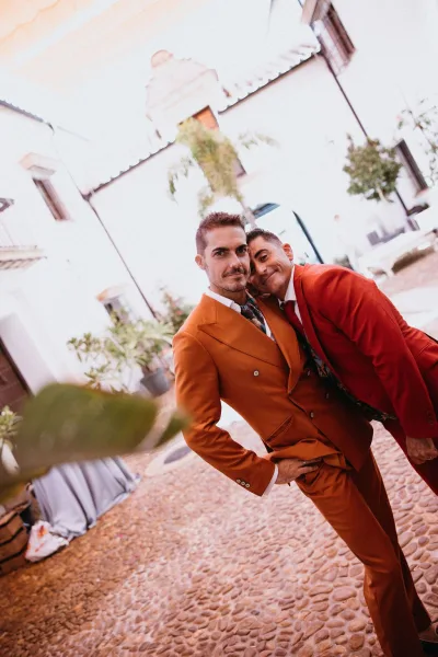 Couple portrait in colorful groom suits—one orange, one red—with neckties, in a cobblestone courtyard with white stucco and potted plants.