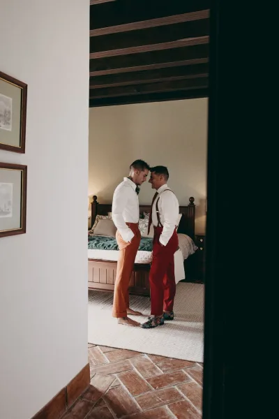 Grooms getting ready share a quiet moment in suspenders in a beamed-ceiling bedroom with a wooden bed and framed art.