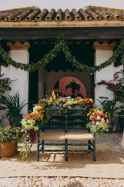 Wedding chair decor with floral chair swags and taper candles in a white stucco courtyard by a clay tile roof and a courtyard archway.