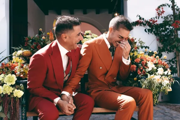 Couple portrait of grooms laughing together on a bench, framed by a white stucco wall and archway with garden vines and cobblestone.