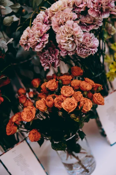 Wedding flowers with orange spray roses arranged in a glass vase on a tabletop, set against blurred foliage and paper cards.