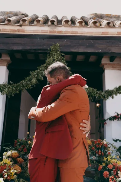 Wedding embrace captured as a candid wedding moment between two grooms at a stucco doorway framed by a greenery garland and wooden beams.