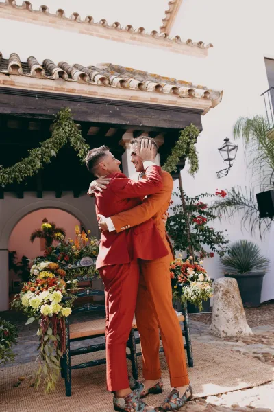 Ceremony moment as grooms embrace beneath an archway, framed by a greenery garland, in a courtyard of white stucco and a clay tile roof.