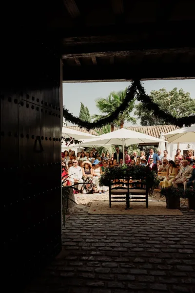 Outdoor ceremony setup for a courtyard wedding ceremony with white umbrellas shading guests in a cobblestone courtyard framed by tall palm trees.