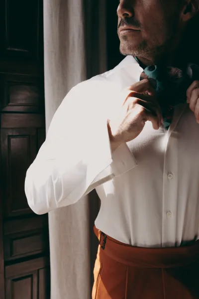Groom getting ready, tying his bow tie as steady hands shape the knot beside a wood door and a curtain.