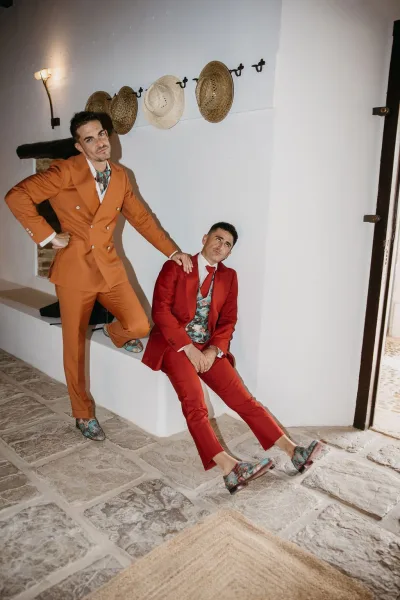 Couple portrait in colorful wedding suits, orange and red, with waistcoats, posed against a white wall with hanging straw hats.