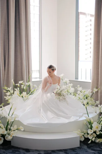 Bridal portrait of a bride holding bouquet of white lilies, seated on a round platform with veil and dress train by large windows overlooking city buildings