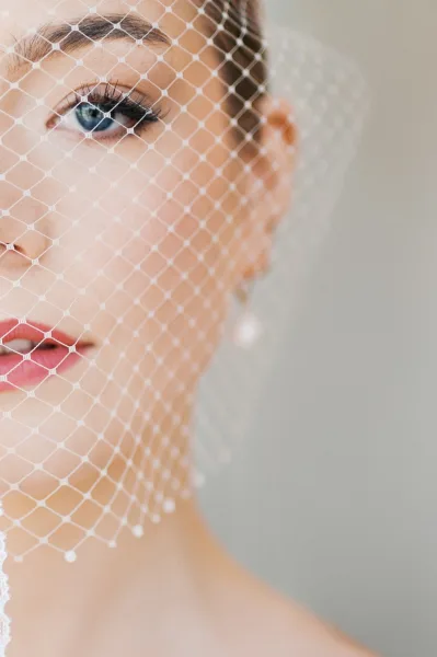 Bridal veil portrait of a bride in a birdcage veil, netting over her face with pearl earring and soft bridal makeup on a neutral backdrop