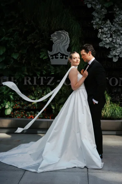 Couple portrait of bride and groom pose, bride facing camera in satin ball gown with long train beside groom in black tux at greenery wall