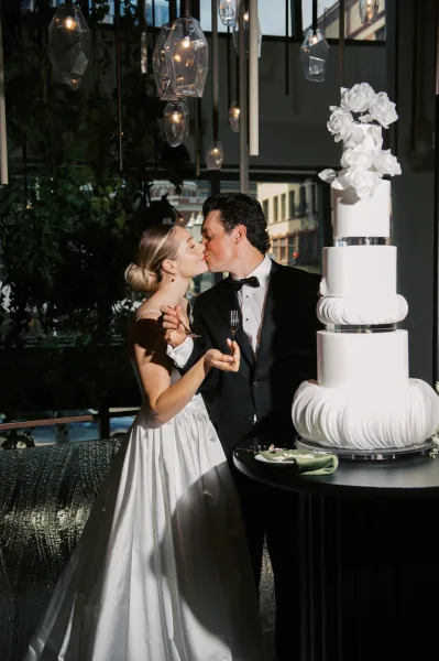 Wedding cake cutting as bride and groom kiss beside a three-tier white cake, champagne flutes and pendant lights glowing by large windows.