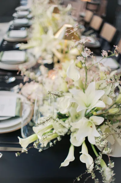 Reception tablescape with wedding table centerpiece of white lilies, bud vases, greenery runner, and candles on a long banquet table indoors