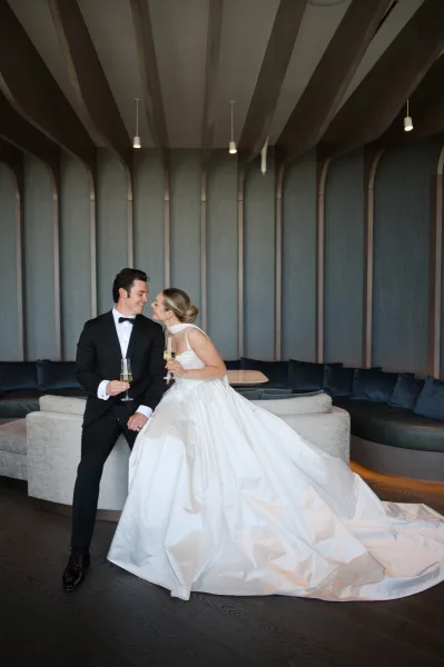 Couple portrait of bride and groom seated on a blue banquette, laughing with champagne flutes in a modern lounge with pendant lights
