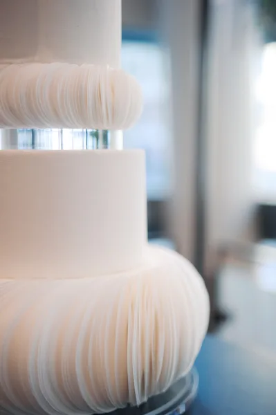 Wedding cake with white wedding cake fondant, tiered design and pleated ruffle texture on a pedestal, softly lit by a window indoors