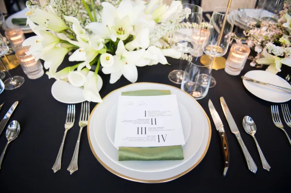 Reception tablescape with wedding place setting on a black tablecloth, gold charger, sage green napkin, menu card, lilies, and ribbed votives