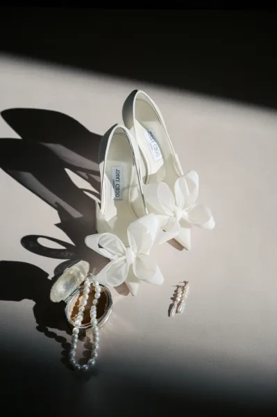 Bridal shoes with white bow wedding heels styled beside a pearl necklace, drop earrings, and ring box on a neutral tabletop in hard light