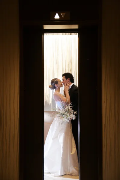 Wedding kiss portrait of bride and groom kissing in an elevator doorway, bride in satin gown holding a calla lily bouquet, earrings glowing in backlight
