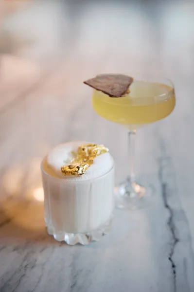 Signature wedding cocktails in cocktail glasses, a foamy coupe with gold leaf garnish and cookie garnish on a marble bar top, bar blurred behind