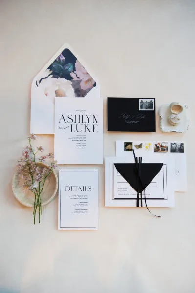 Invitation suite wedding invitation flat lay with floral envelope liner, black RSVP envelope, stamps, wax seal, ribbon, and rings on neutral surface