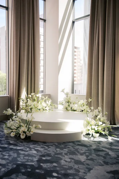 Wedding ceremony backdrop with a round white stage and calla lily floral accents, set before floor-to-ceiling windows with sunlit city views
