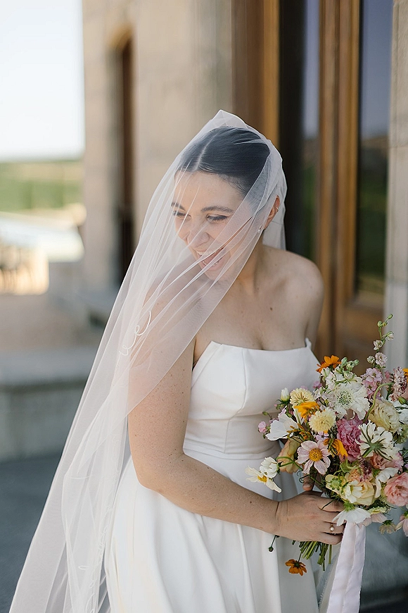 Bridal portrait of a bride with veil, holding a colorful bridal bouquet and showing her engagement ring by a stone building walkway