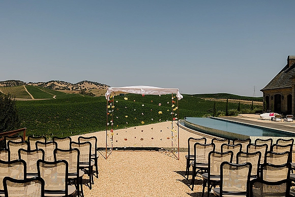Ceremony setup with outdoor ceremony seating facing a draped floral arch by a reflecting pool in a gravel courtyard with vineyard hills