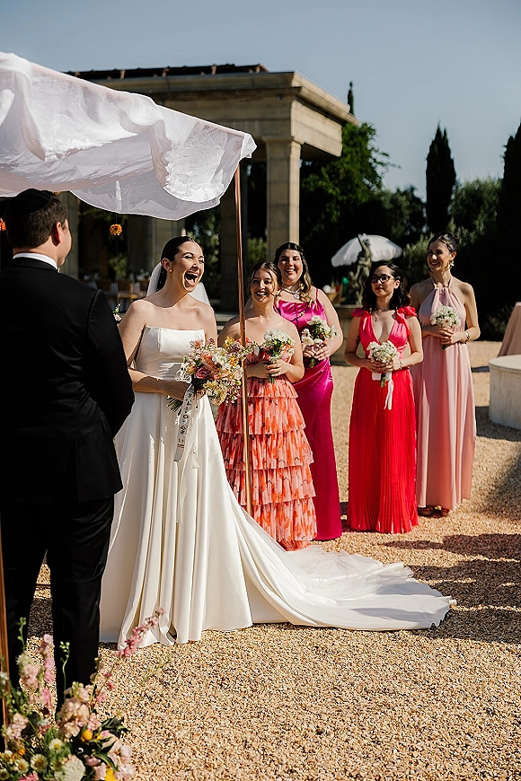 Ceremony moment under a wedding chuppah canopy as bride laughs in strapless dress and veil, groom in tux, bridesmaids in bright dresses outdoors