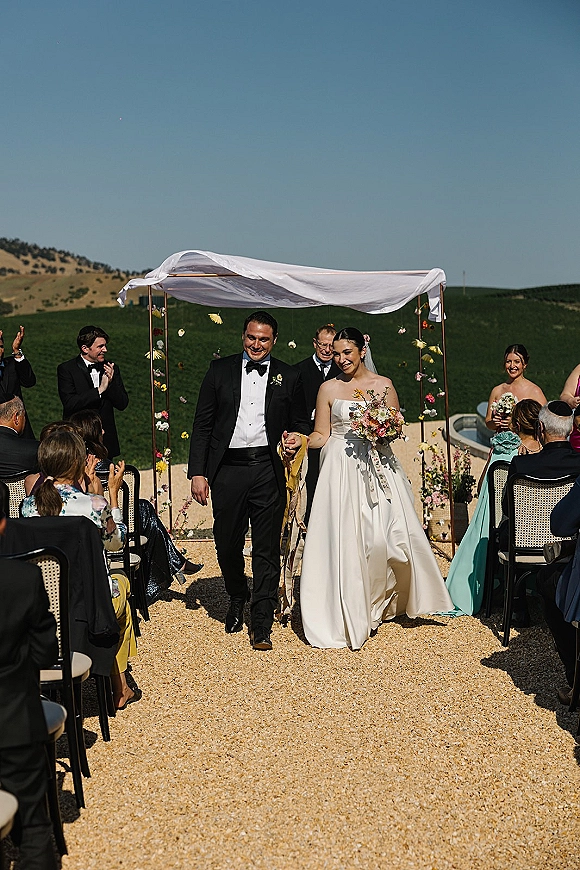 Wedding recessional as bride and groom walk the aisle under a draped floral arch, guests clapping beside gravel path with green hills beyond