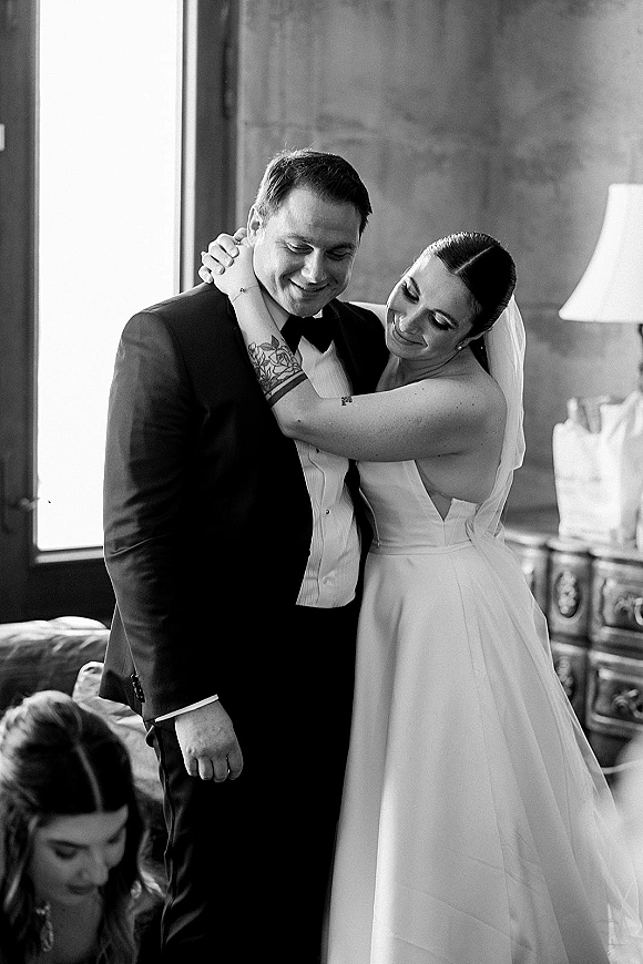 Couple portrait in black and white wedding portrait style, bride in veil hugging groom in tuxedo by window light beside sofa and lamp