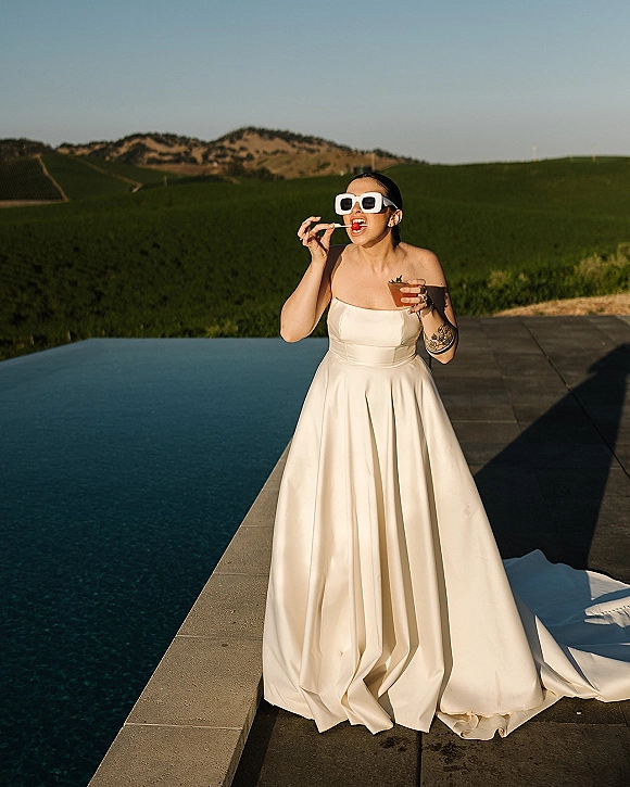 Bridal portrait of a bride in sunglasses sipping a cocktail in a strapless wedding dress by an infinity pool with green hills beyond