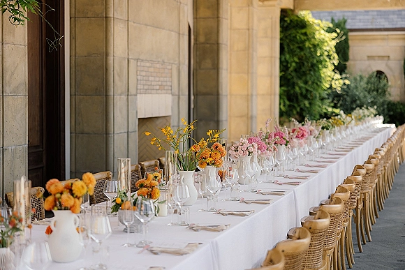 Reception tablescape with a long banquet table, white linens, pink and orange flowers in ceramic vases, and rattan chairs in a stone courtyard colonnade