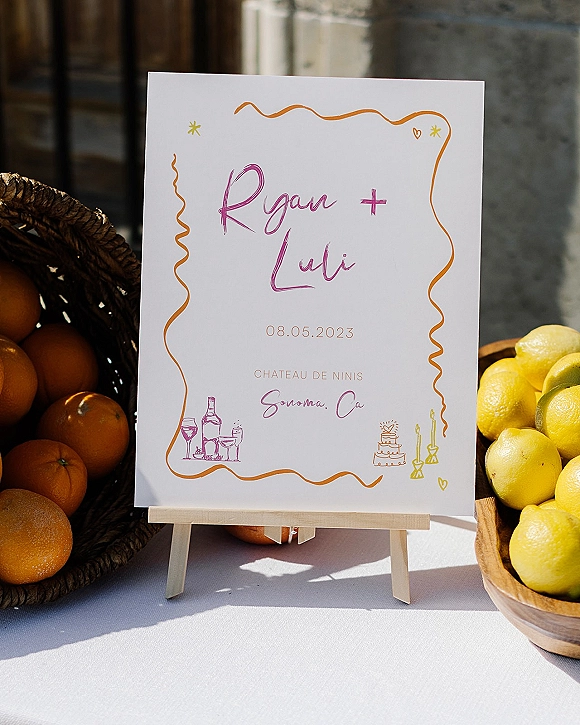 Wedding welcome sign with hand lettering on a small easel, accented by citrus fruit in bowls and basket on a linen table by a stone wall