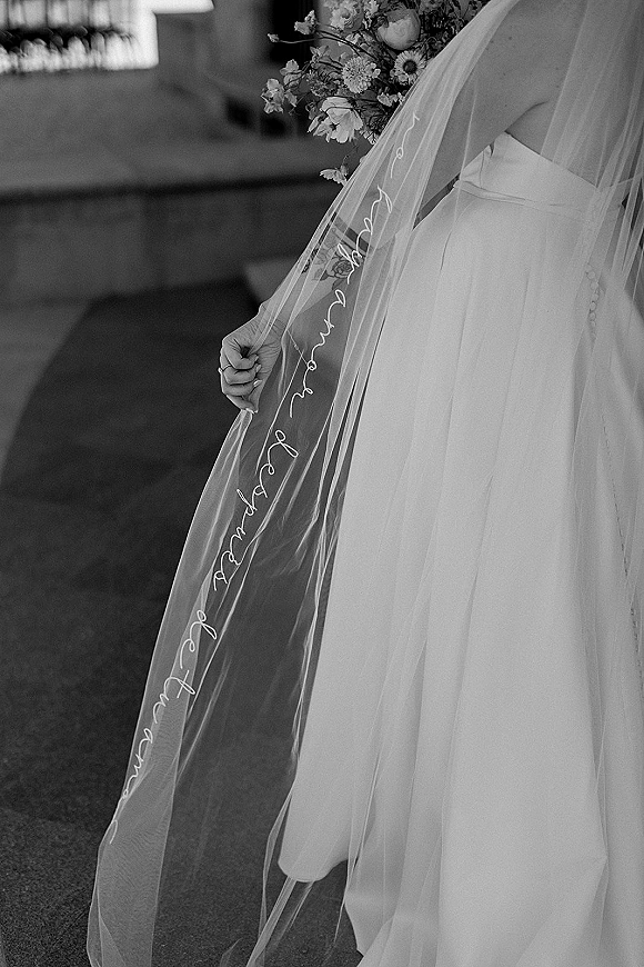 Bridal veil detail with embroidered wedding veil lettering draped over a bouquet and ring, with church pews softly blurred behind