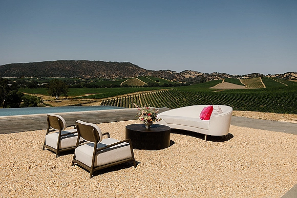Lounge seating decor with a white chaise, modern chairs and round coffee table, accented by a floral arrangement on a poolside vineyard patio
