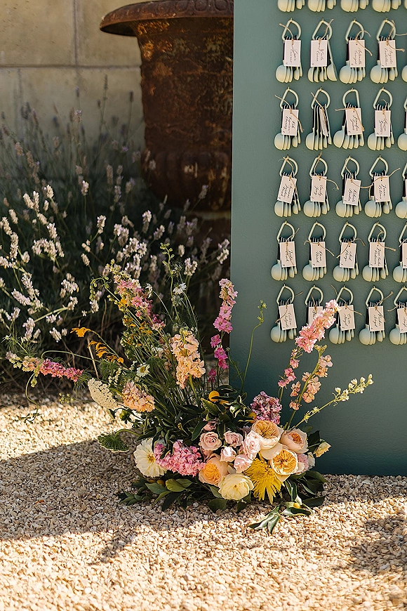 Escort card display with wedding escort cards on teal board, hanging calligraphy tags and mini scoops, framed by wildflower florals near lavender bushes