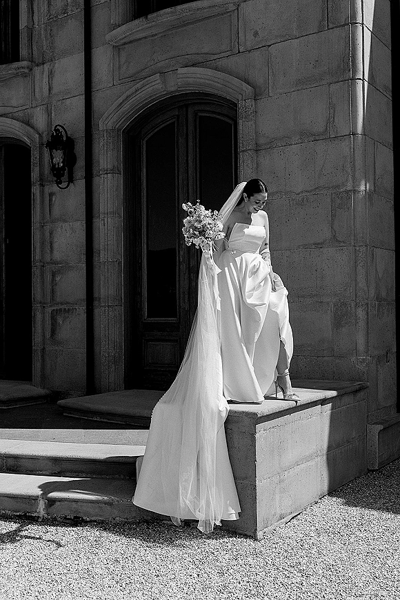 Bridal portrait in black and white of a bride holding bouquet, wearing a strapless dress and long veil on steps by a stone archway