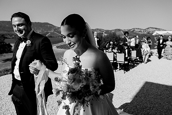 Wedding recessional as bride and groom walk hand in hand down a gravel aisle, bride holding bouquet with long ribbons, guests and hills behind